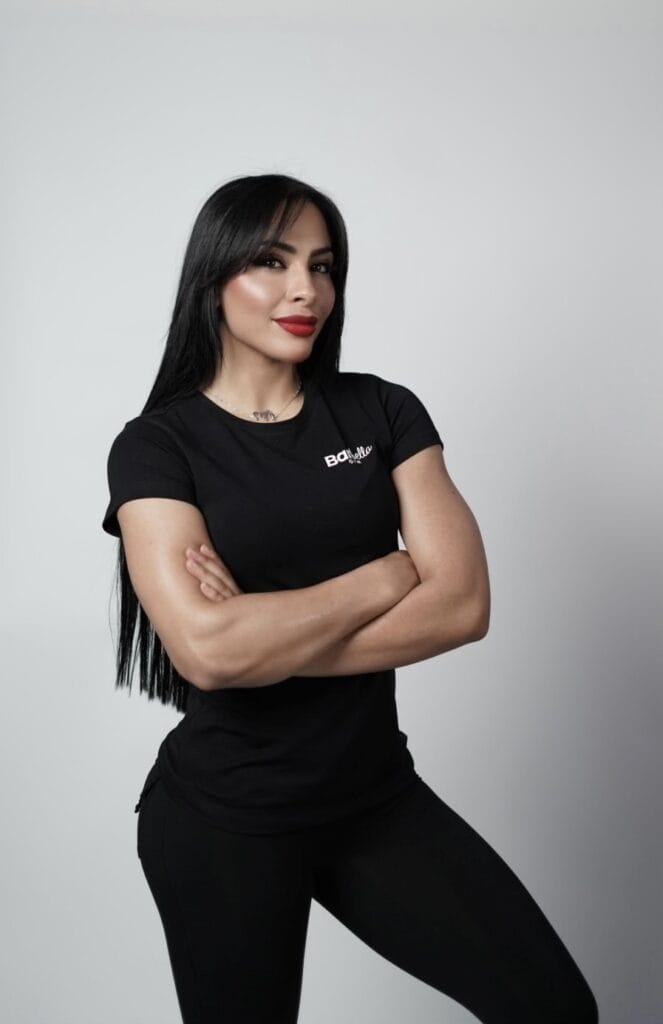 A woman with long black hair and red lipstick stands against a plain light gray background. She is wearing a fitted black T-shirt and black pants, crossing her arms and smiling slightly. The T-shirt has a small white logo on the chest that reads "Botlife.