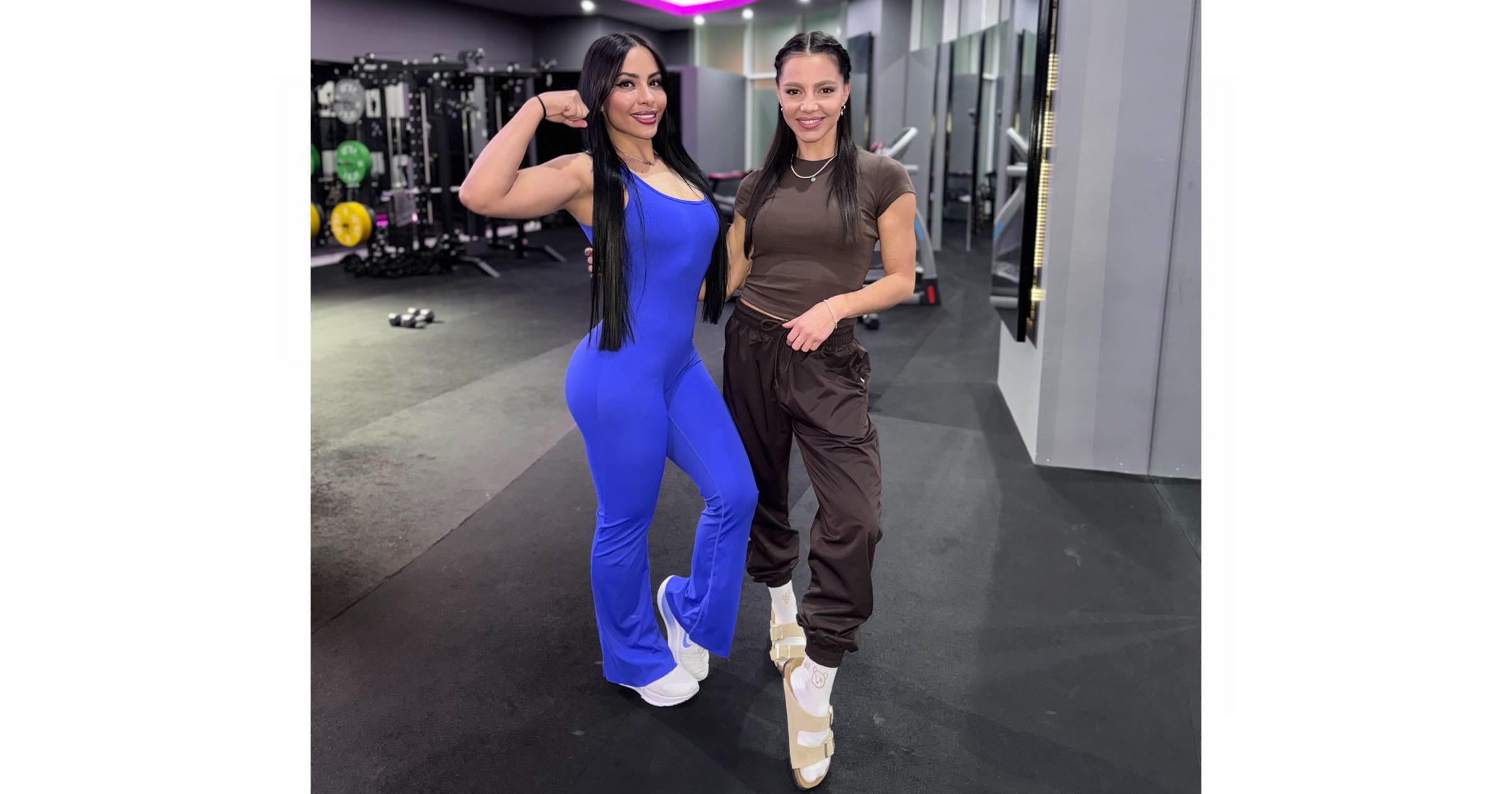 Two women pose in a gym. One flexes her arm and wears a bright blue jumpsuit; the other smiles and wears brown pants, a brown t-shirt, and white sneakers. Gym equipment and weights are visible in the background under bright lighting.